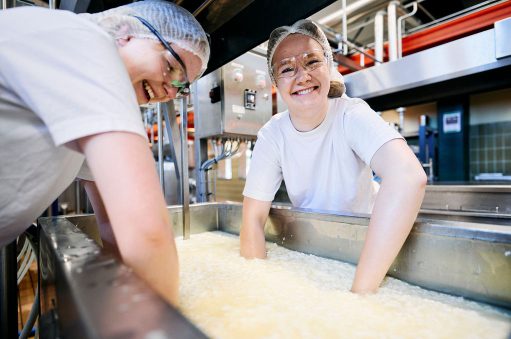 To smilende elever arbejder med ostemasse i et mejerianlæg, iført beskyttende hårnet og briller.